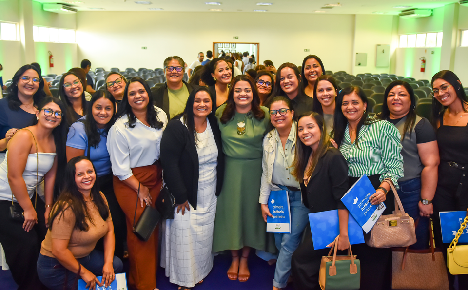 Grupo de mulheres reunidas em um auditório, sorrindo, com pastas da campanha Agosto Verde nas mãos