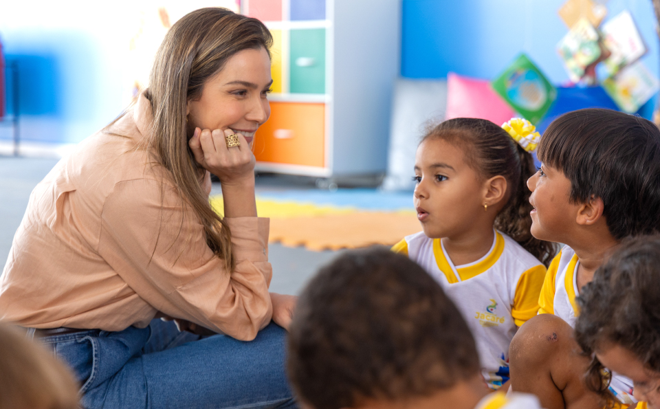 Mulher sentada no chão de uma sala de educação infantil conversa com crianças de uniforme escolar